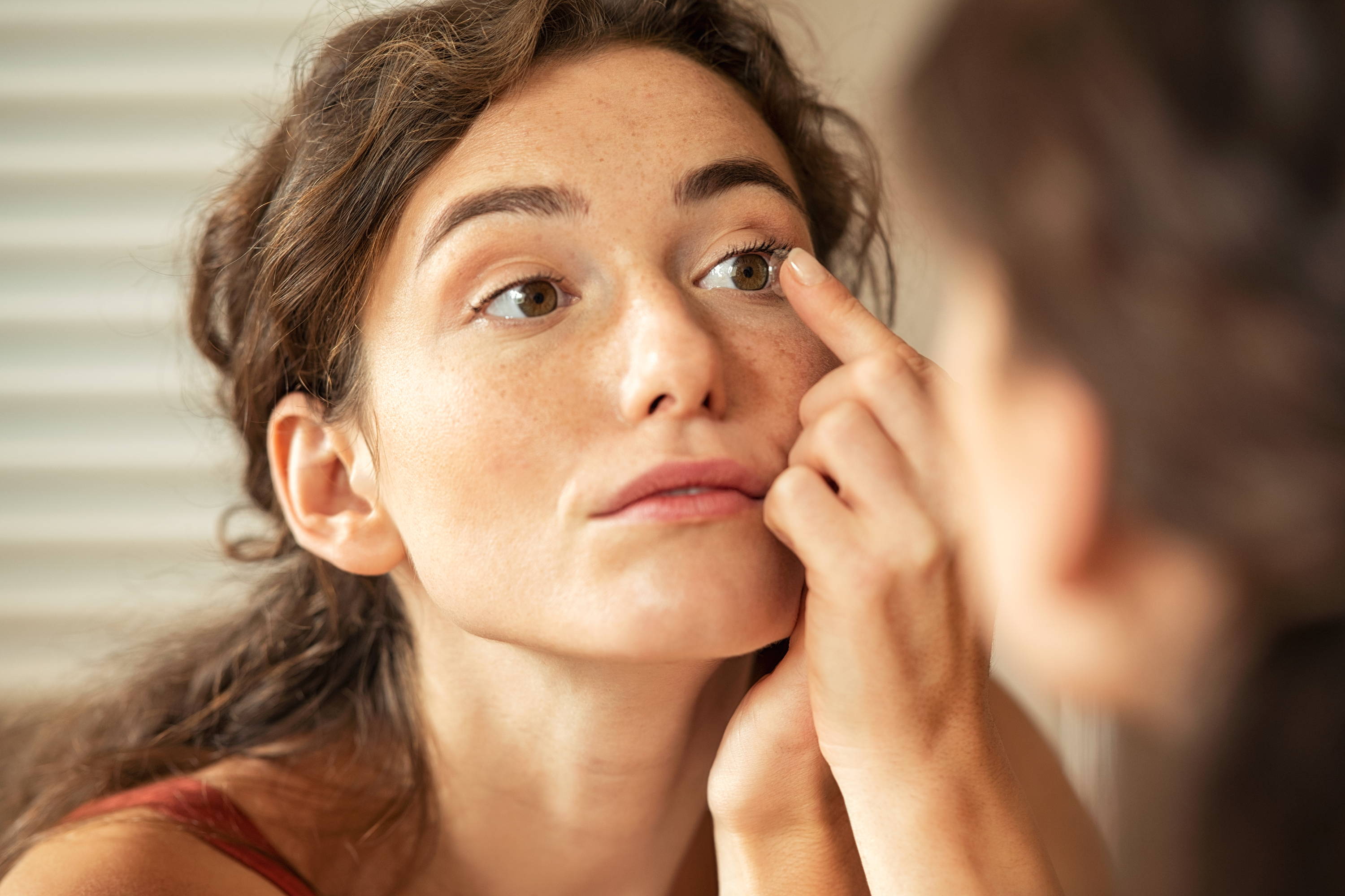 Una mujer joven se mira al espejo mientras cuidadosamente se coloca un lente de contacto.