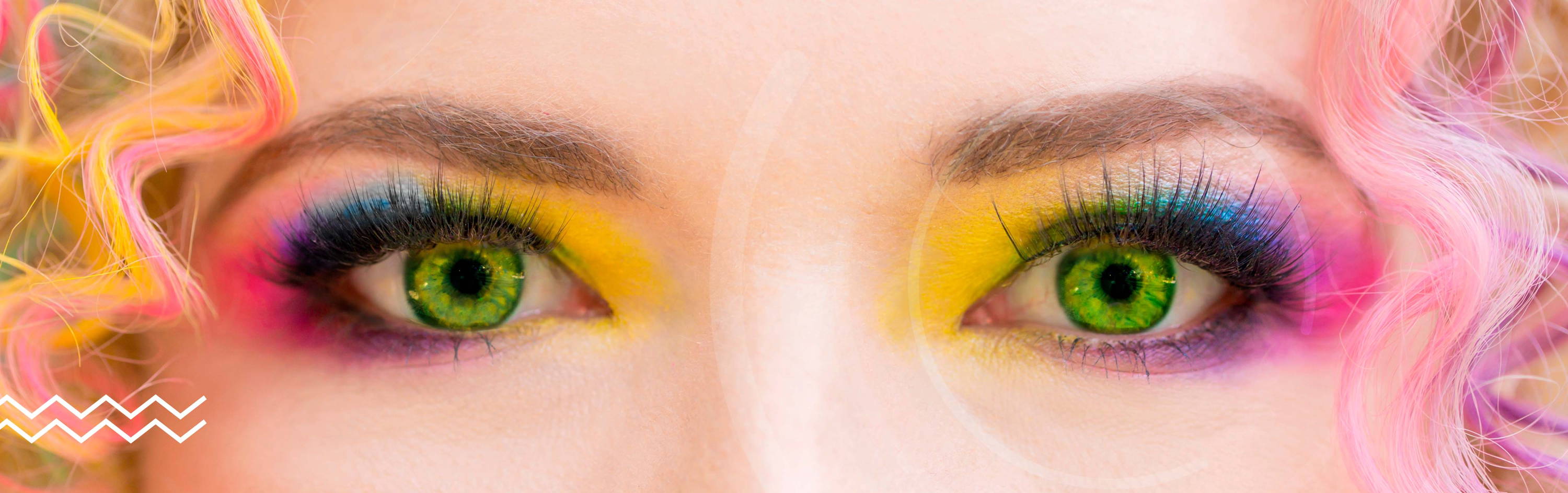 Imagen de los ojos de una mujer que lleva puesto unos lentes de contacto de color en tono verde. Aunque la imagen principal son los ojos de la modelo, podemos apreciar que trae una peluca y maquillaje con los colores del arcoiris.