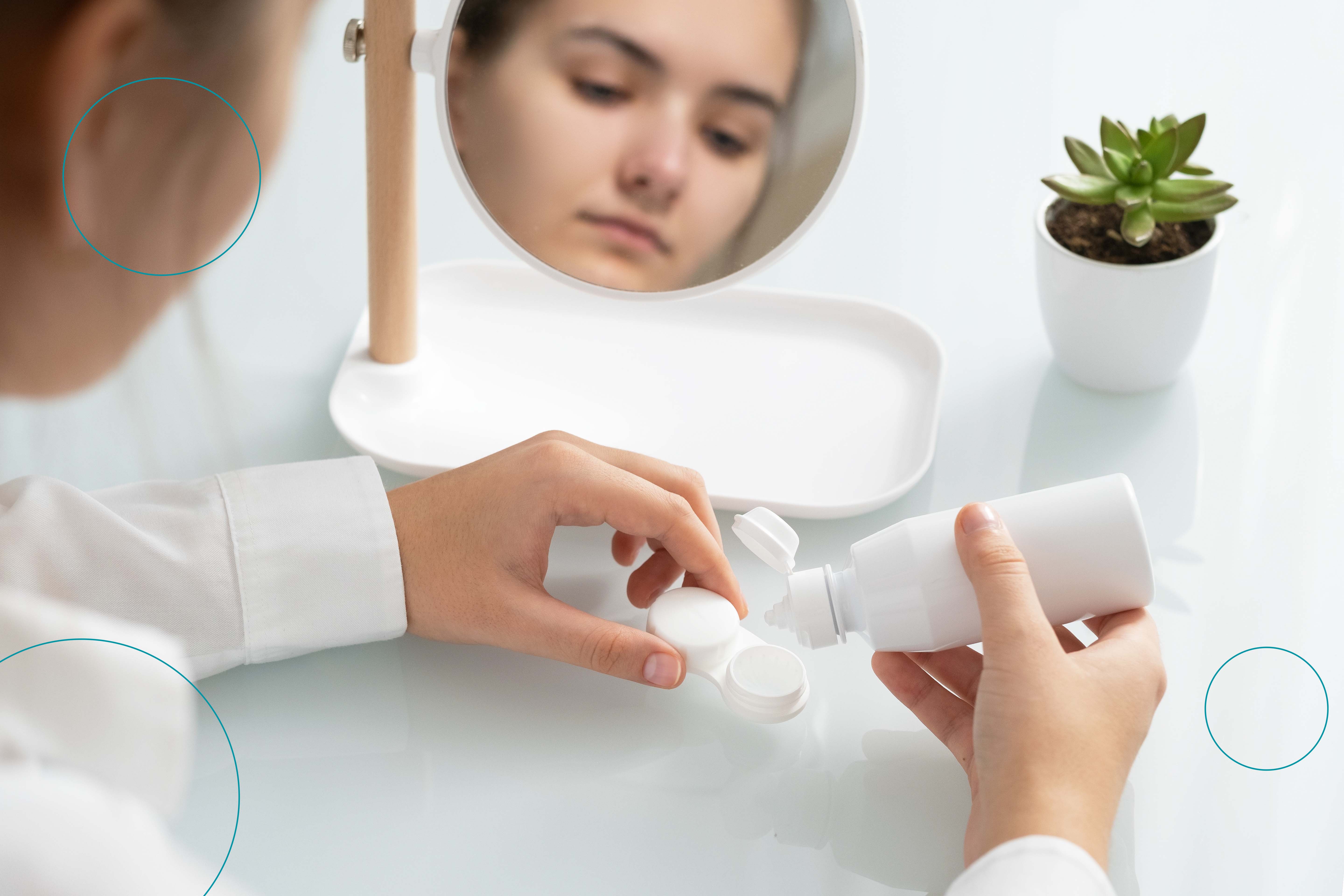 Una mujer en un escritorio frente a un espejo con una planta, vaciando líquido para limpiar sus lentes con contacto 